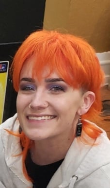 Portrait of a smiling person with bright orange hair wearing a light gray hoodie and black shirt