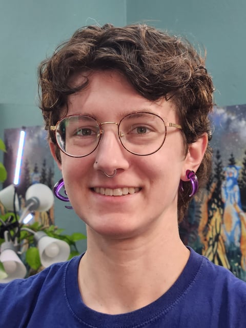 Smiling person with short brown wavy hair wearing round glasses and purple hoop earrings, navy shirt, against blurred indoor background