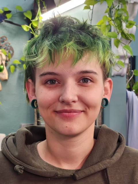 Person with short dyed green hair, black hoop earrings, and a brown hoodie smiling at the camera indoors with hanging plants behind them