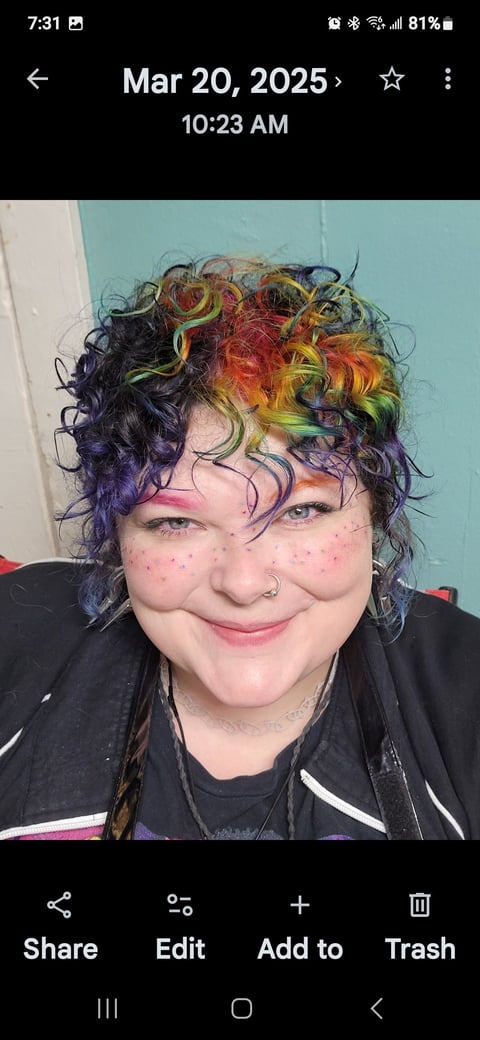 Person with colorful rainbow curly hair and nose piercing smiling at camera against turquoise background