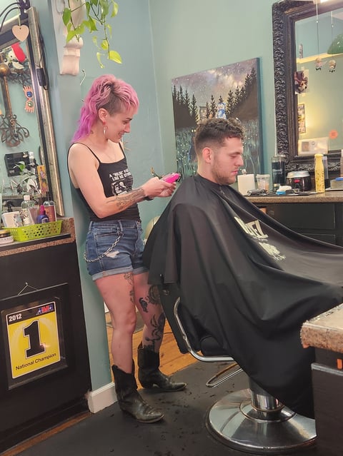 Hairstylist with pink hair cutting a male clients hair in a salon with teal walls and decorated with plants and artwork