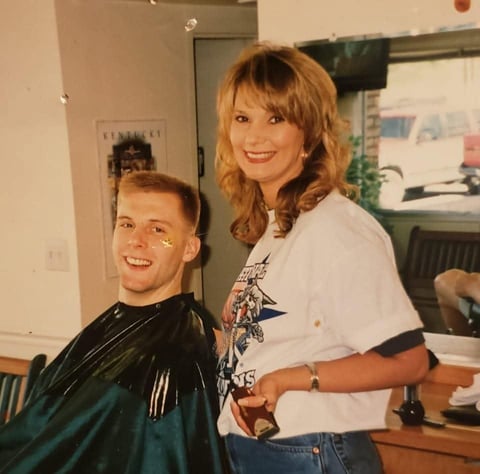 Woman with reddish-blonde hair standing behind a young man in a barber cape, smiling in an indoor salon setting