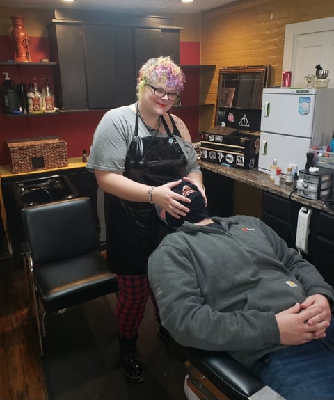 Tattoo artist with purple hair wearing glasses and apron works on clients arm in studio